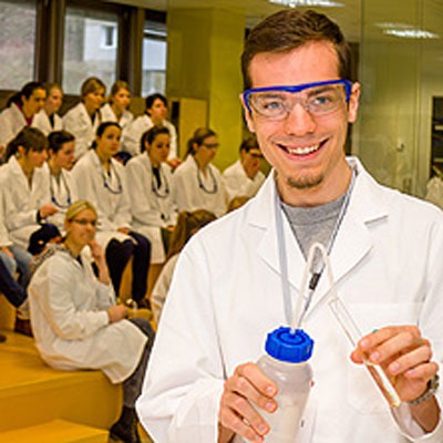 Explorer - 9. bis 12. Schulstufe=Junger Laborant demonstriert Experiment, im Vordergrund einer Schulklasse