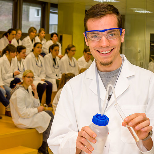 Explorer - 9. bis 12. Schulstufe=Junger Laborant demonstriert Experiment, im Vordergrund einer Schulklasse